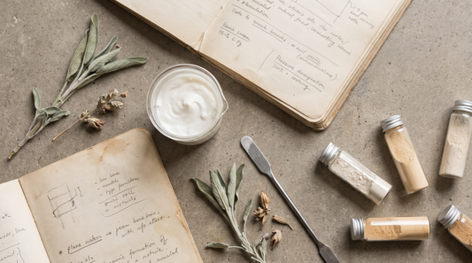 A formulation workspace with a beaker of cream, glass vials of powder, a spatula, a notebook and a dried botanical sprig.